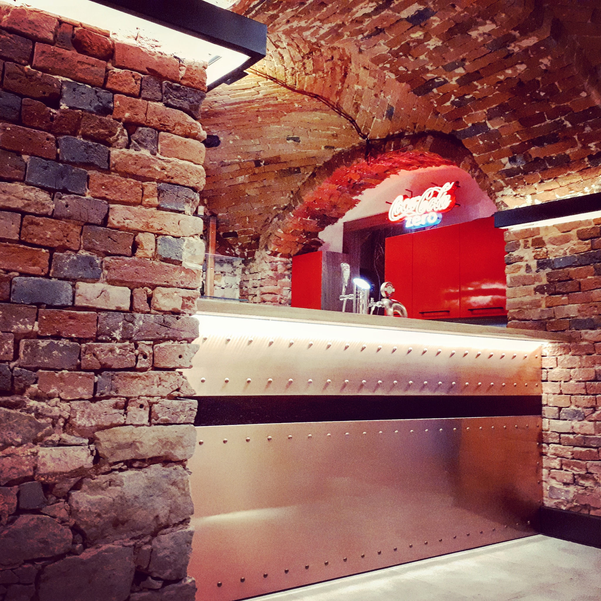 Bar z blachy z nitami, podświetlony od góry, w pomieszczeniu z ceglanym sklepieniem łukowym i neonem Coca-Cola Zero w tle.