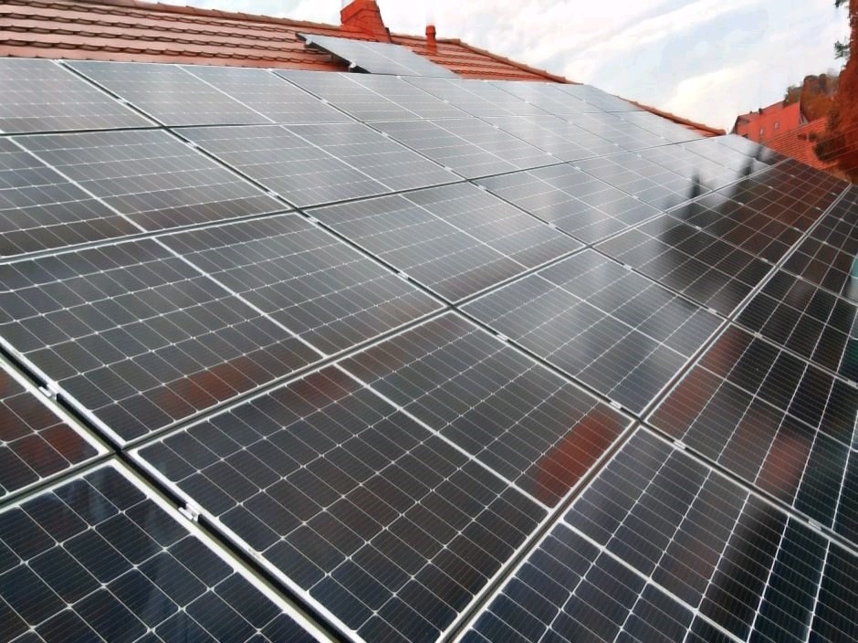 Panele fotowoltaiczne zamontowane na dachu pokrytym czerwoną dachówką, widok z dołu pod kątem, częściowo zachmurzone niebo w tle.