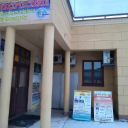 Wejście do biura ubezpieczeń z szyldem 'Ubezpieczenia w jednym miejscu, 20 Towarzystw' i tablicami informacyjnymi różnych firm ubezpieczeniowych, w tym oferta ubezpieczeń na życie i OC/AC.