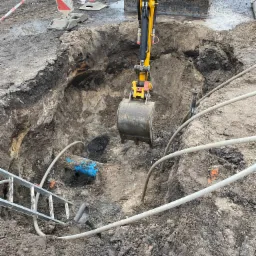 Żółta mini koparka nad głębokim wykopem w ziemi, widoczne rury, drabina i oznaczenia ZWiK.