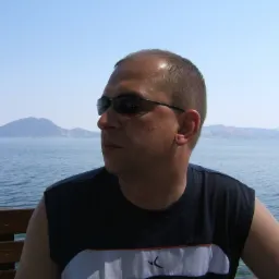 Mężczyzna w okularach przeciwsłonecznych siedzi na ławce z widokiem na morze i górzysty krajobraz w tle.