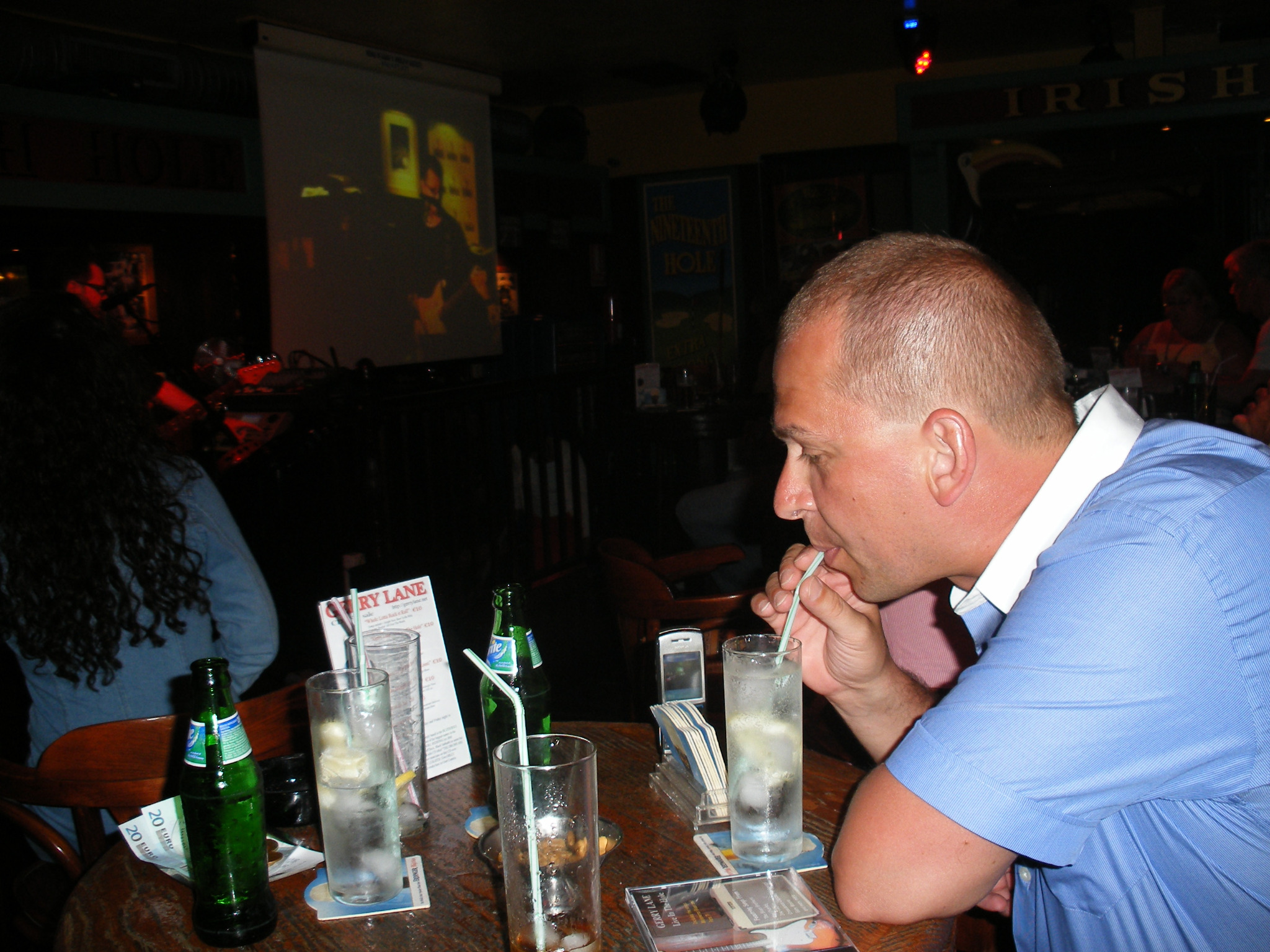 Mężczyzna w niebieskiej koszuli pije drinka ze słomką w barze, na stole stoją szklanki, butelka piwa i menu z cenami w euro, w tle wyświetlany jest film na projektorze.