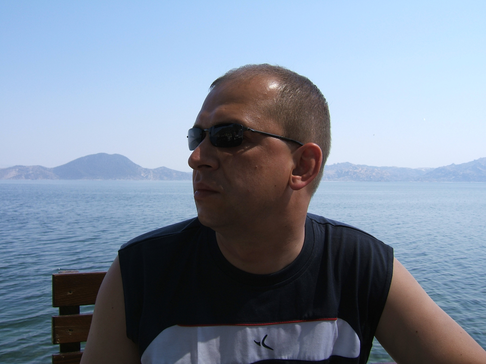 Mężczyzna w okularach przeciwsłonecznych siedzi na ławce z widokiem na morze i górzysty krajobraz w tle.