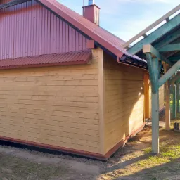 Nowo wybudowany drewniany budynek z czerwonym dachem z blachy falistej i konstrukcją drewnianej pergoli w trakcie budowy, widoczne świeżo pomalowane na zielono słupy i belki.