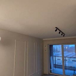 Arttiler Glazurnictwo Marcin Józefowicz - Wnętrze pokoju po remoncie z dekoracyjnymi listwami na ścianie, nowoczesną lampą wiszącą z kulistymi kloszami i reflektorami sufitowymi, widokiem na miasto z balkonu.