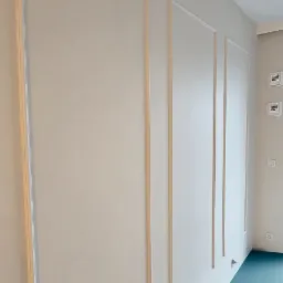 Ściana z dekoracyjnymi listwami w kolorze naturalnego drewna, tworzącymi prostokątne panele, na tle białej ściany i turkusowej podłogi.