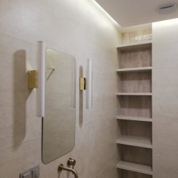 Arttiler Glazurnictwo Marcin Józefowicz - Beżowa łazienka z podświetlanym lustrem, złotą armaturą i wnęką z półkami wyłożoną pionową mozaiką. Minimalistyczny design, ciepłe oświetlenie.