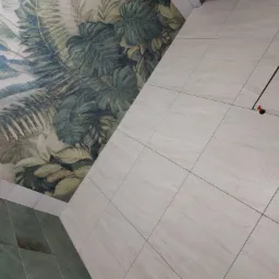 Układanie beżowych płytek podłogowych z motywem liści na ścianie, widoczne systemy poziomowania płytek.