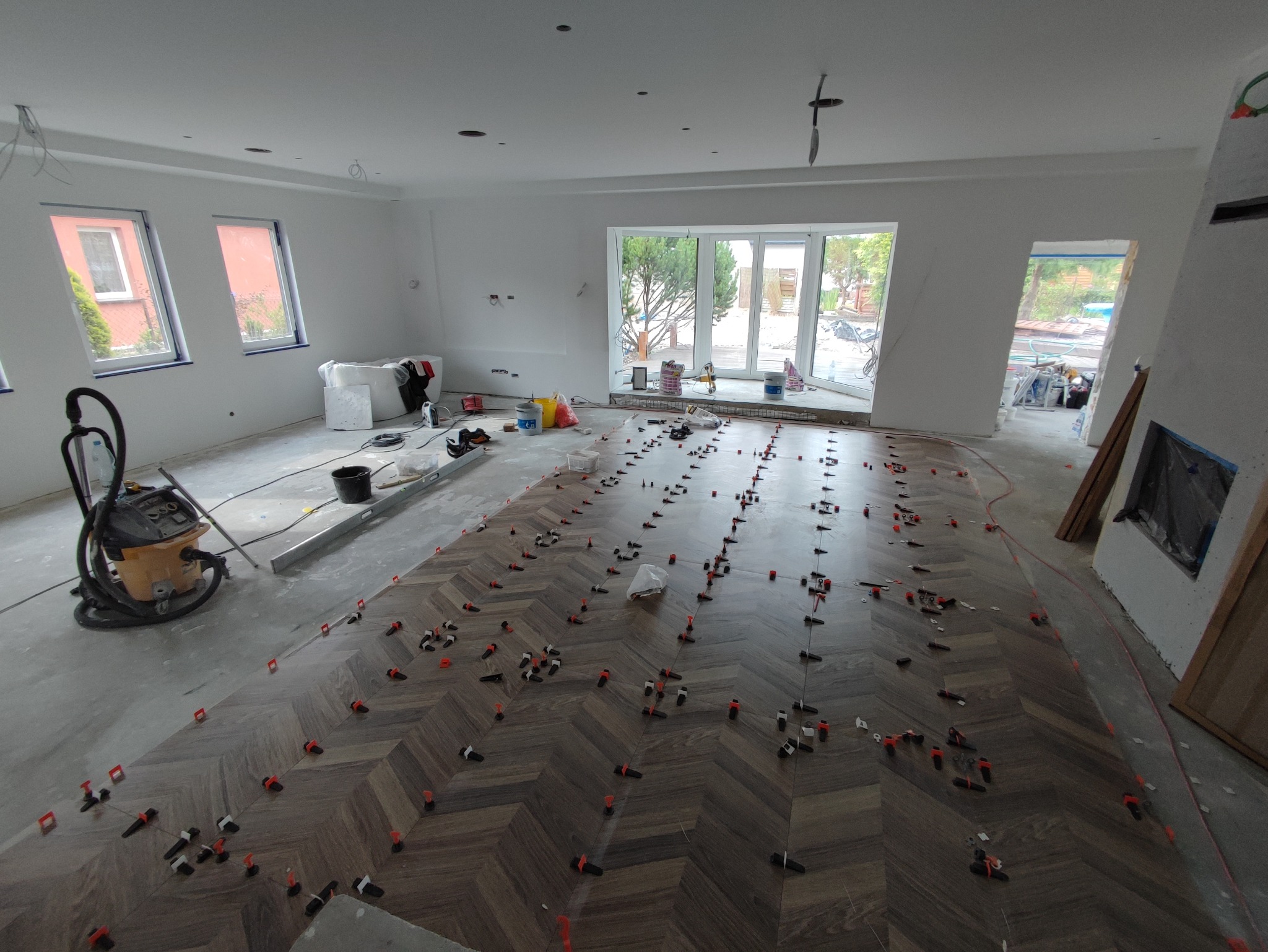 Układanie parkietu w jodełkę w przestronnym, jasnym pomieszczeniu; widoczne narzędzia, poziomica, odkurzacz przemysłowy i system poziomowania płytek.