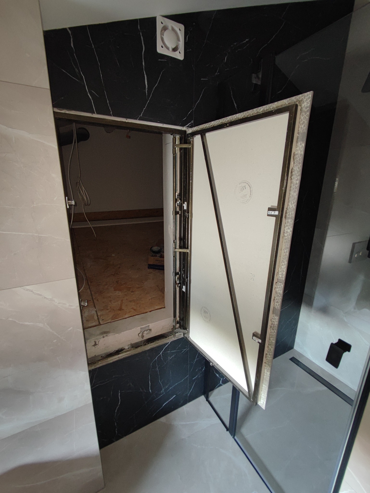 Otwarty rewizyjny właz podtynkowy w łazience z czarno-białymi marmurkowymi płytkami, widoczne przewody i podłoże wewnątrz.