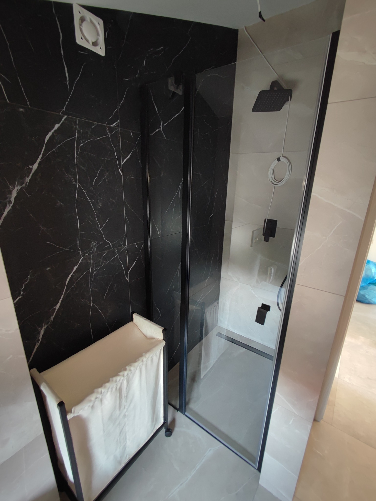 Nowoczesna łazienka z kabiną prysznicową typu walk-in, wyłożona płytkami imitującymi czarny marmur z białymi żyłami oraz jasnymi płytkami na pozostałych ścianach, z widocznym koszem na pranie w rogu.