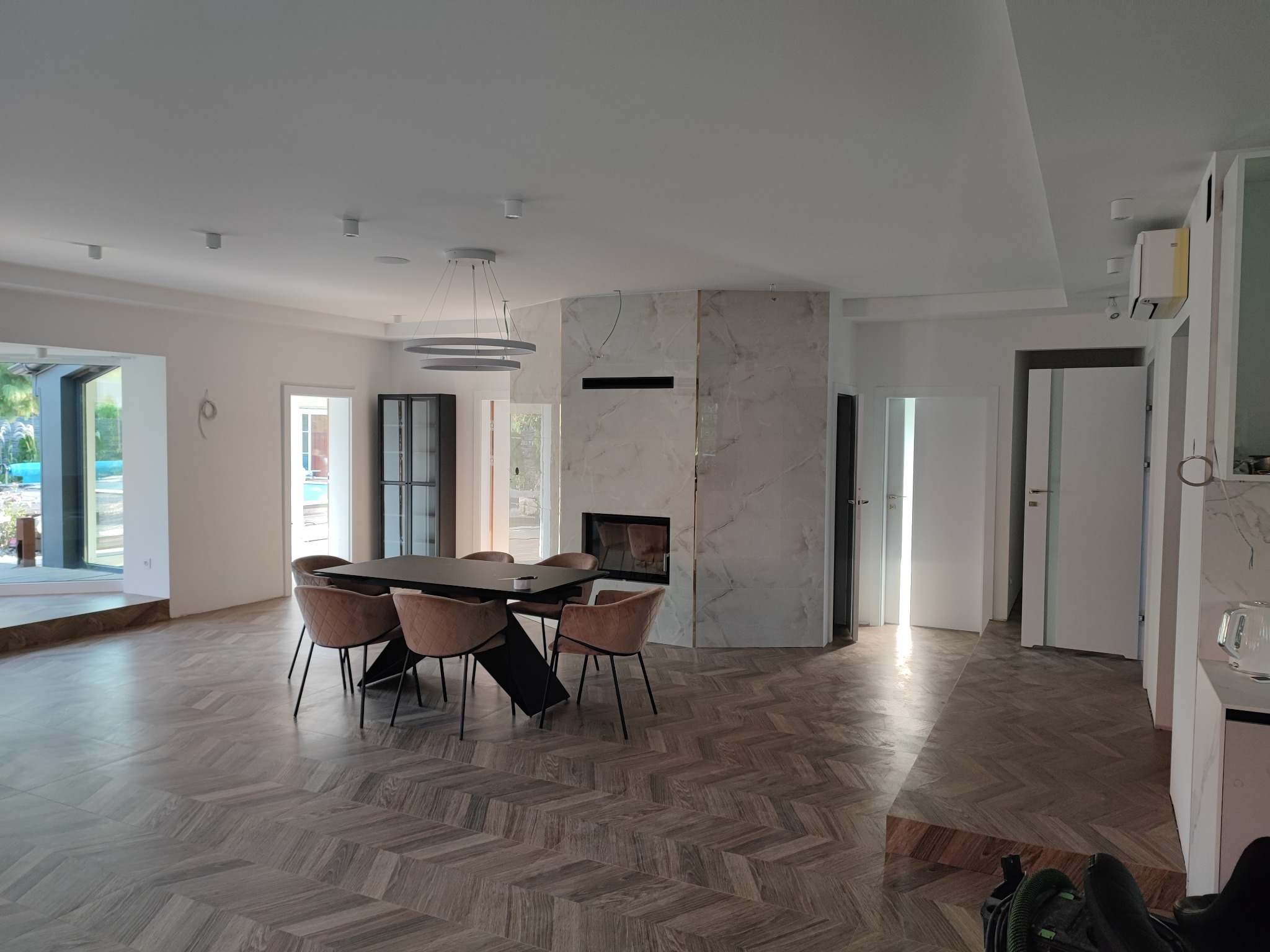 Przestronny salon z parkietem w jodełkę, marmurowym kominkiem i nowoczesnym oświetleniem, stół z krzesłami. Widoczne drzwi i okna. W rogu odkurzacz przemysłowy.