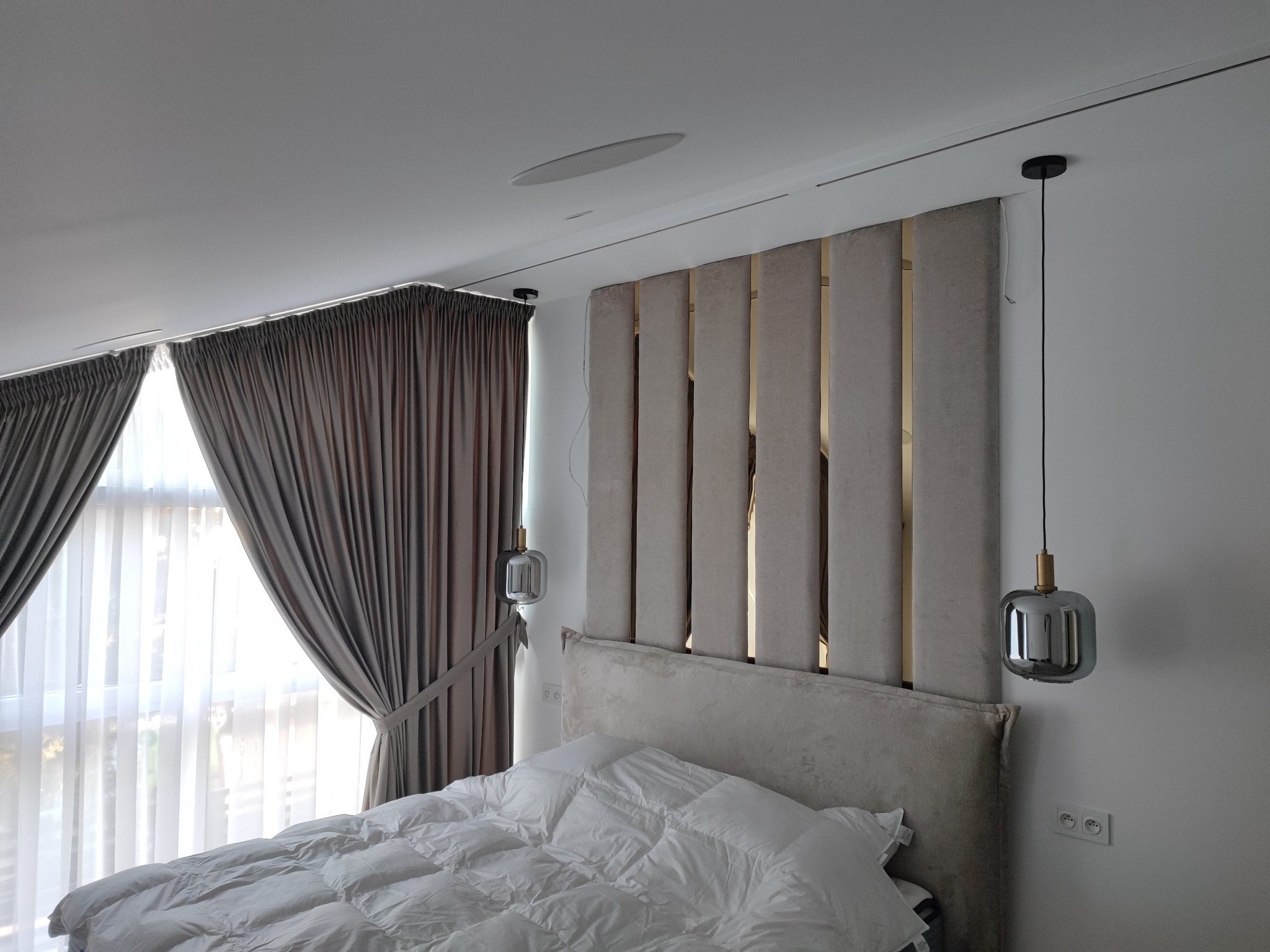 Sypialnia z beżowym tapicerowanym panelem za łóżkiem, szarymi zasłonami i nowoczesnymi lampami wiszącymi.