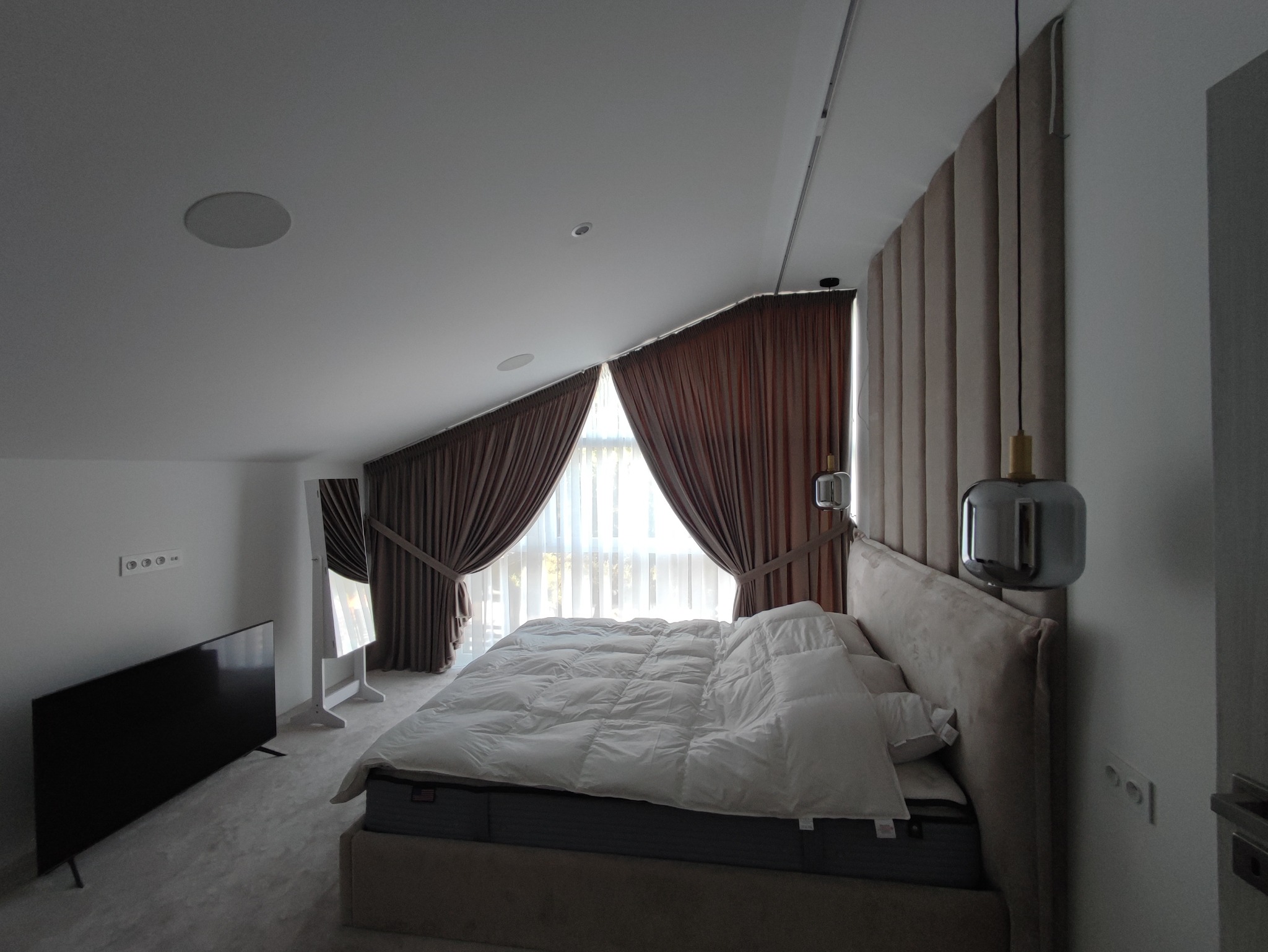 Nowoczesna sypialnia na poddaszu ze skośnym sufitem, brązowymi zasłonami, panelem tapicerowanym za łóżkiem i minimalistyczną lampką nocną.