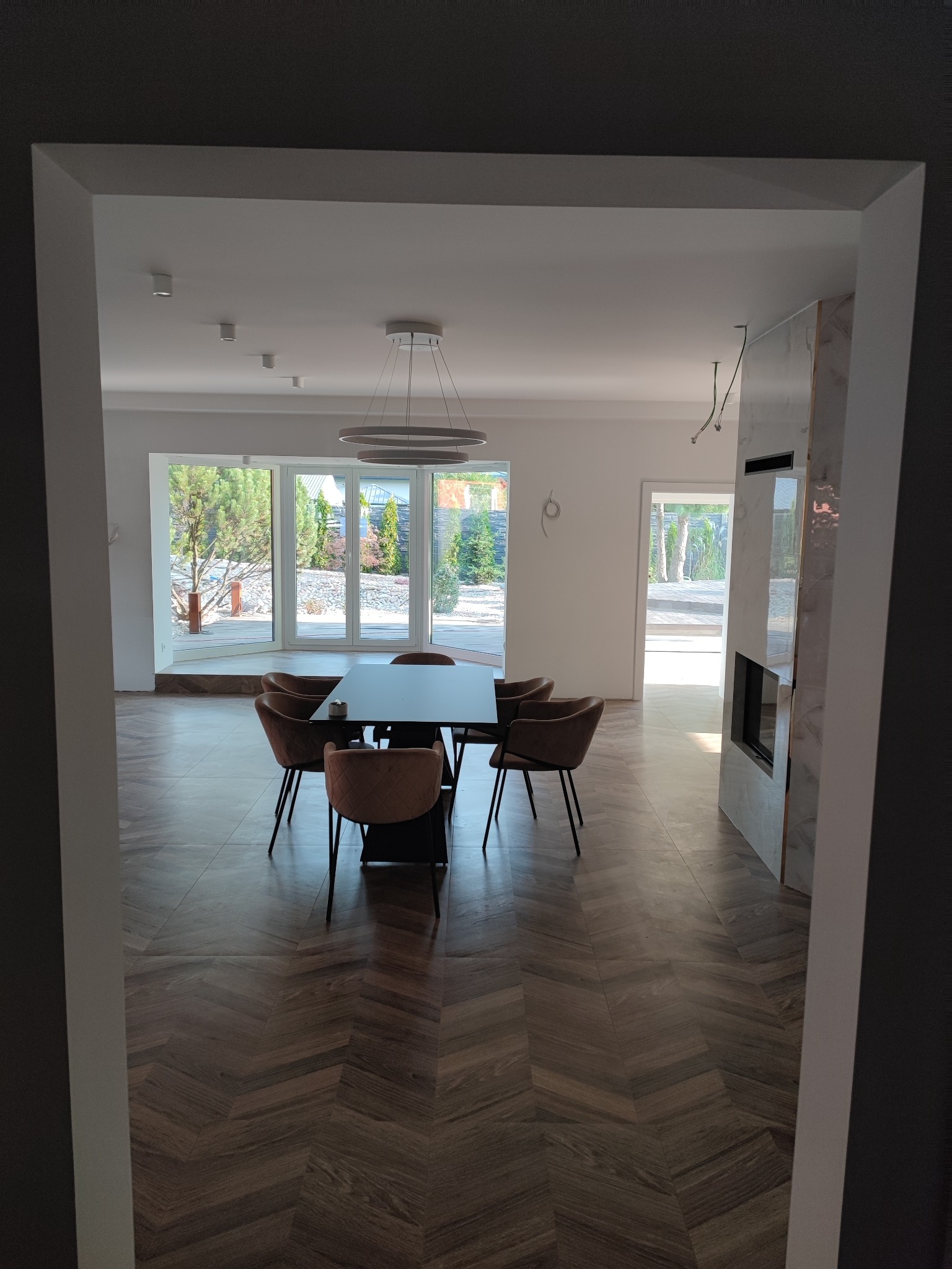 Nowoczesna jadalnia z ciemną podłogą ułożoną w jodełkę, czarnym stołem i brązowymi krzesłami, widok na ogród przez panoramiczne okna, minimalistyczny kominek obłożony marmurem.