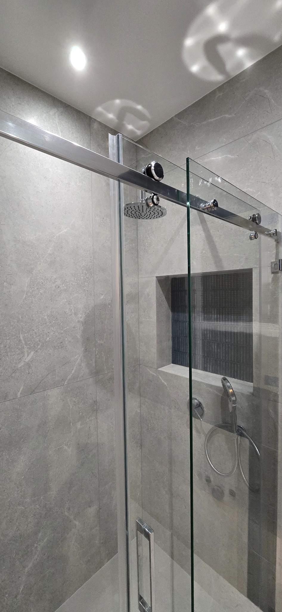 Szklana kabina prysznicowa z przesuwnymi drzwiami, chromowanymi okuciami i deszczownicą, zamontowana na ścianie wyłożonej szarymi płytkami imitującymi marmur, z wnęką wykończoną ciemnymi płytkami.
