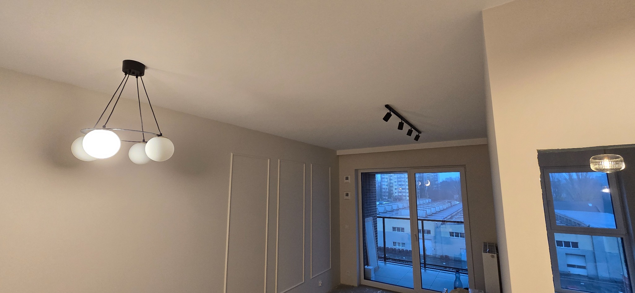 Wnętrze pokoju po remoncie z dekoracyjnymi listwami na ścianie, nowoczesną lampą wiszącą z kulistymi kloszami i reflektorami sufitowymi, widokiem na miasto z balkonu.