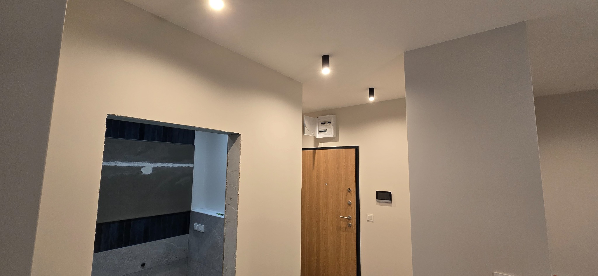 Wnętrze mieszkania w trakcie remontu: widoczny fragment łazienki z ułożonymi płytkami, surowy otwór drzwiowy oraz drzwi wejściowe z panelem sterowania i skrzynką elektryczną pod sufitem.