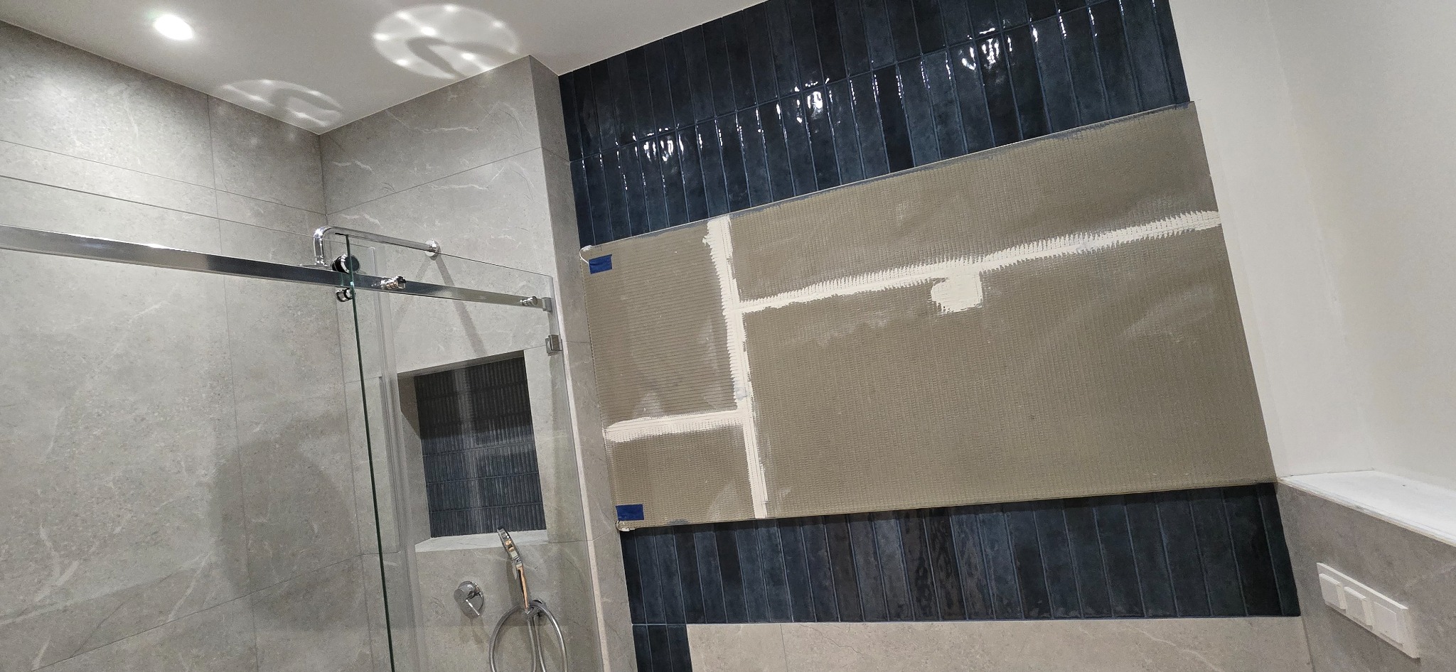 Montaż ciemnych, pionowych płytek w łazience, widoczna siatka montażowa i fragment kabiny prysznicowej z szarymi płytkami.