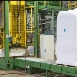 Wielki, biały, zapakowany i zabezpieczony ładunek na palecie, umieszczony na zielonym przenośniku taśmowym w hali produkcyjnej z żółto-zieloną konstrukcją maszyny w tle.