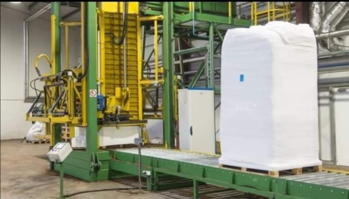 Wielki, biały, zapakowany i zabezpieczony ładunek na palecie, umieszczony na zielonym przenośniku taśmowym w hali produkcyjnej z żółto-zieloną konstrukcją maszyny w tle.