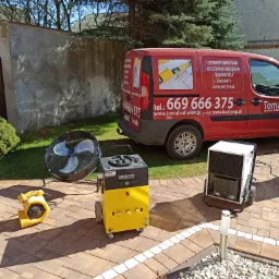 Sprzęt do osuszania: wentylator, nagrzewnica i osuszacze powietrza, ustawione na kostce brukowej przed budynkiem i czerwonym samochodem firmy TomaBud z reklamą usług osuszania mieszkań.