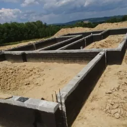 Fundamenty domu jednorodzinnego w trakcie budowy, ściany fundamentowe z bloczków betonowych zabezpieczone izolacją pionową, widoczne pręty zbrojeniowe, tło stanowią drzewa i pagórkowaty teren...