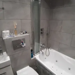 Nowoczesna łazienka w szarościach z wanną z hydromasażem, kabiną prysznicową i podwieszaną toaletą. Na ścianach płytki imitujące kamień, minimalistyczna armatura i akcesoria.