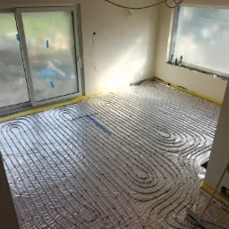 Rozłożona instalacja ogrzewania podłogowego na folii z siatką, widoczne rury w kształcie pętli, okna zabezpieczone folią malarską, przygotowanie do wylewki.