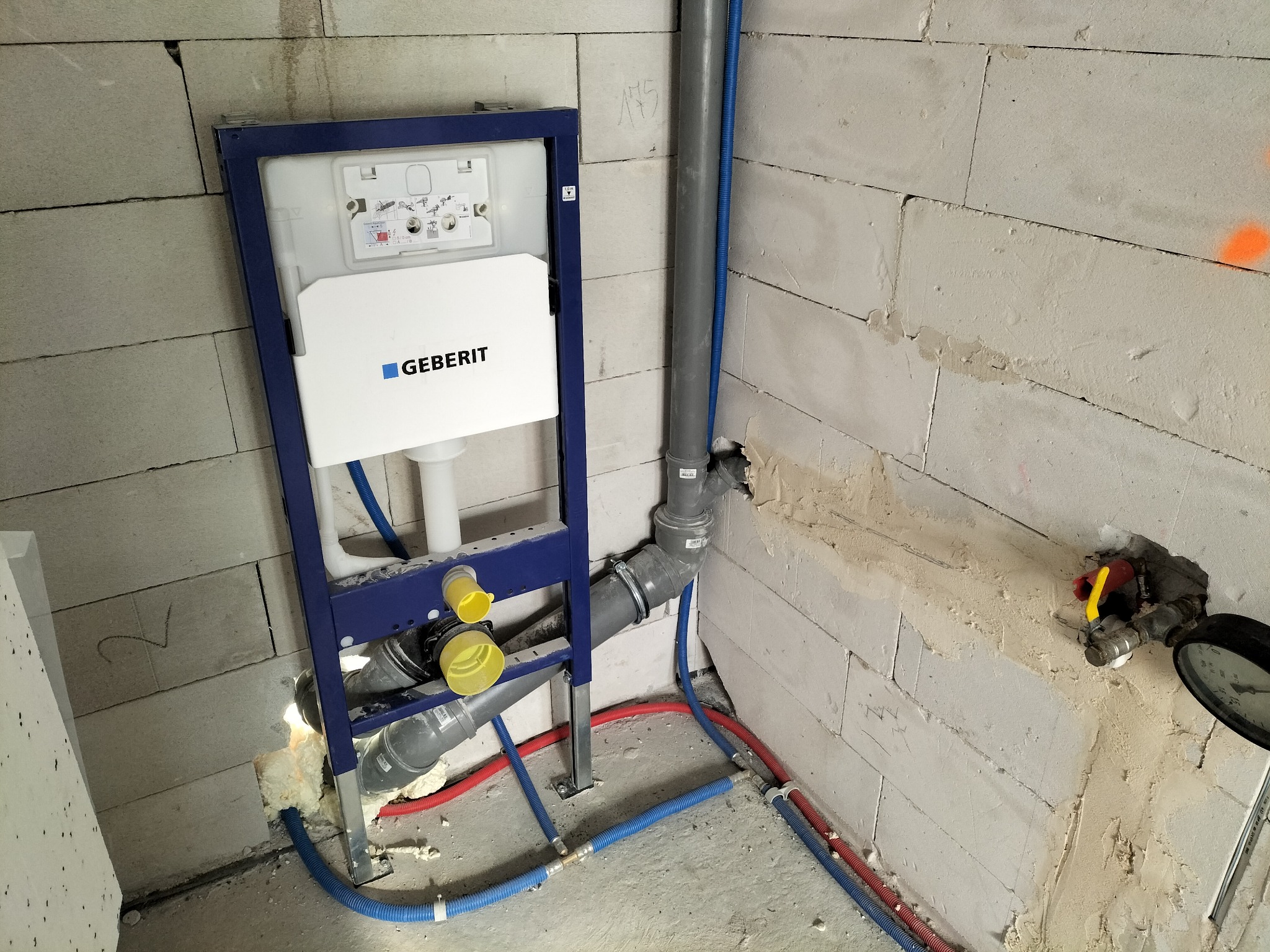 Stelaż podtynkowy Geberit zamontowany przy surowej ścianie z betonu komórkowego, widoczne rury kanalizacyjne i instalacje wodne w kolorze niebieskim i czerwonym, manometr zamontowany na ścianie.