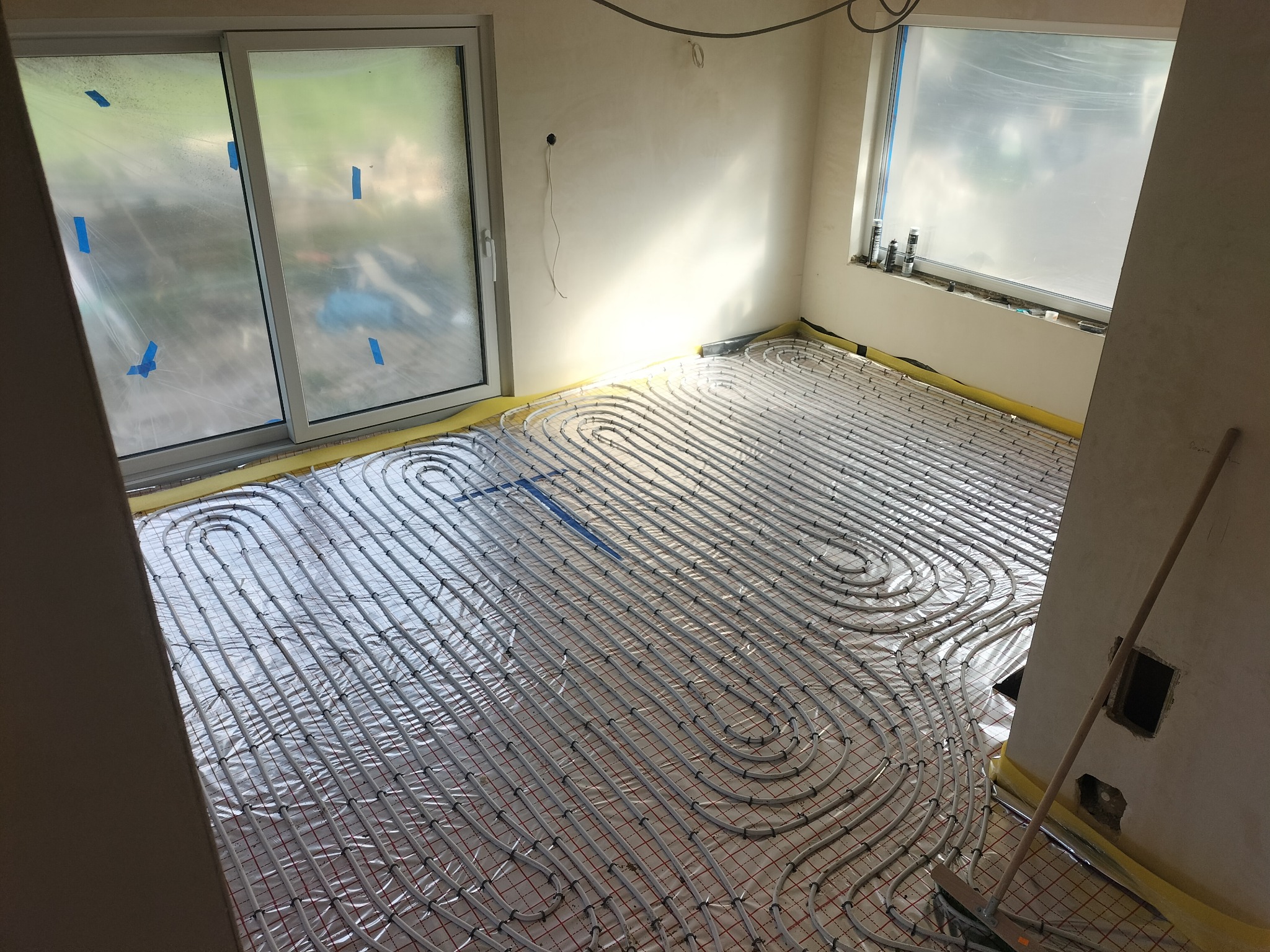 Rozłożona instalacja ogrzewania podłogowego na folii z siatką, widoczne rury w kształcie pętli, okna zabezpieczone folią malarską, przygotowanie do wylewki.