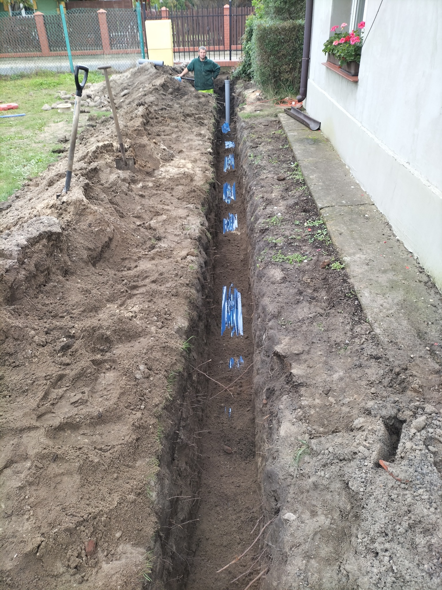 Wykop pod instalację wodną z rurami zabezpieczonymi niebieską folią, widoczny pracownik i łopaty na hałdzie ziemi.