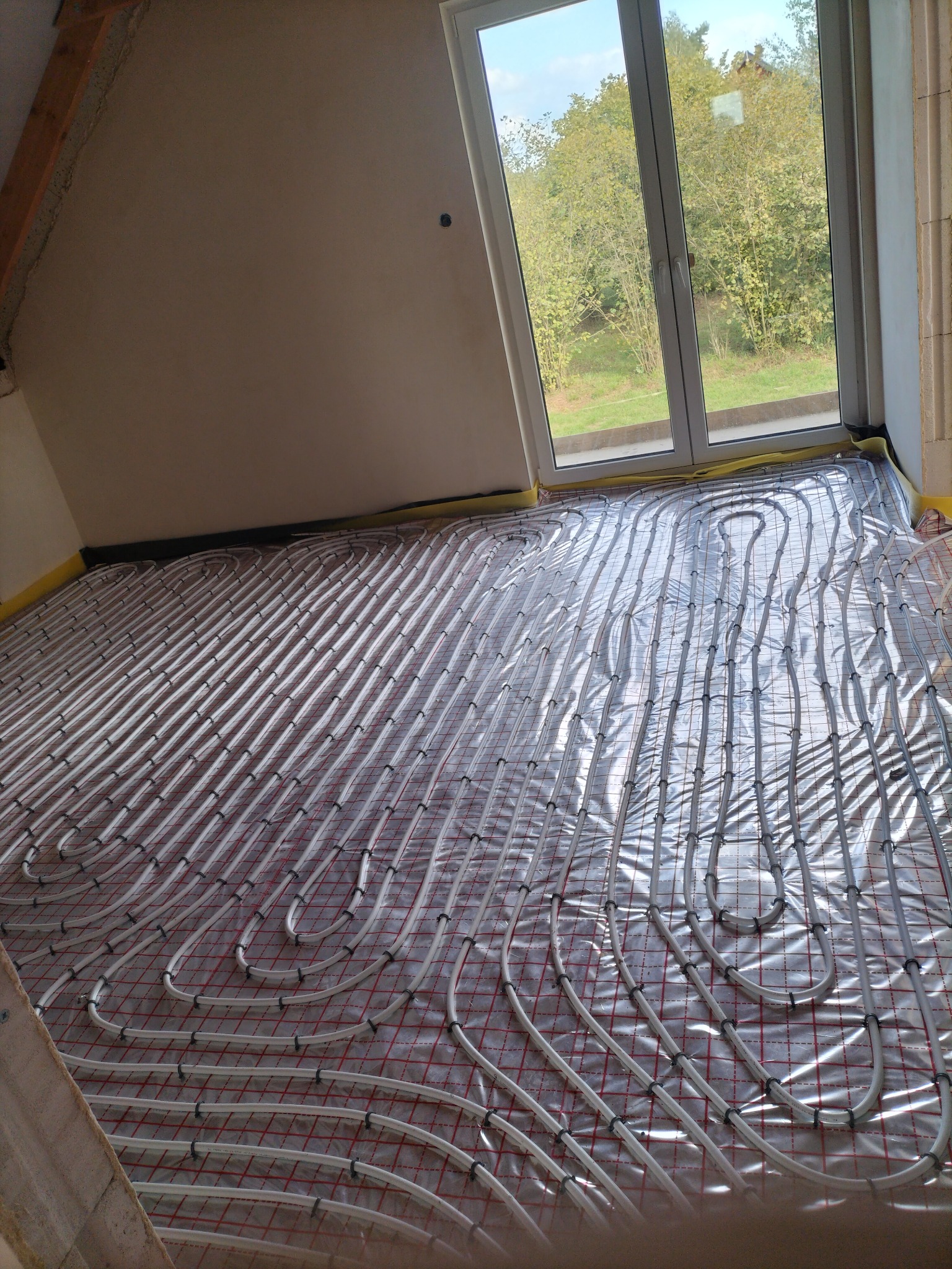 Białe rurki ogrzewania podłogowego ułożone na czerwonej siatce w nowym pomieszczeniu z oknem wychodzącym na zieleń.