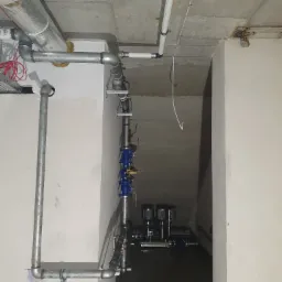 Surowy betonowy sufit z instalacjami: rury stalowe ocynkowane, rury PCV, przewody elektryczne. W tle widoczny system pompowy z dwoma pompami.