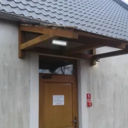 Szary dach z blachy trapezowej nad wejściem do budynku z drewnianą wiatą i oświetleniem LED, widoczny komin i drzewa w tle.