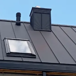 Szary dach z blachy na rąbek stojący z oknem dachowym, kominem wentylacyjnym i ozdobnym kominem. Widok na błękitne niebo.