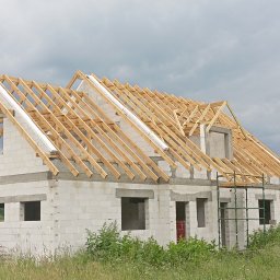 Usługi dekarsko-ciesielskie Ireneusz Rożek - Wznoszenie drewnianej więźby dachowej na murowanym domu z betonu komórkowego. Widoczne białe membrany dachowe i rusztowanie. W tle pochmurne niebo.