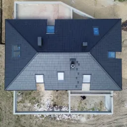 Widok z góry na dwukolorowy dach z dachówki Brass Turmalin (szary i grafitowy) z oknami dachowymi i kominami na surowym terenie budowy z fundamentami.