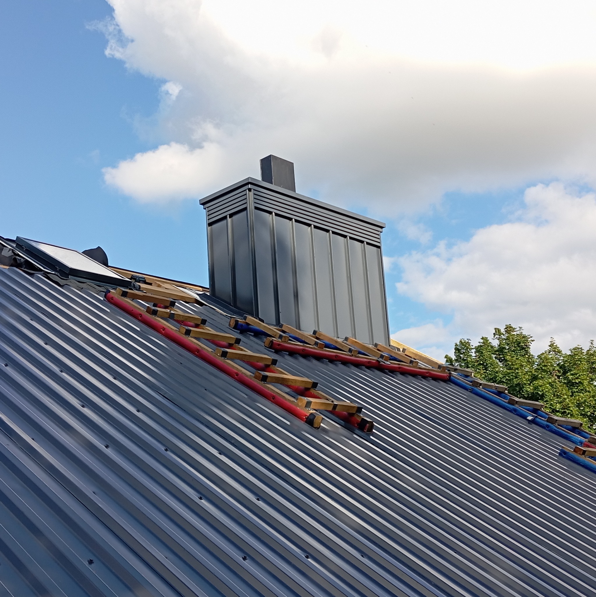 Szary dach z blachy trapezowej w trakcie montażu, z drabinkami dekarskimi i kominem na tle błękitnego nieba z chmurami. Widoczne elementy mocujące blachę.