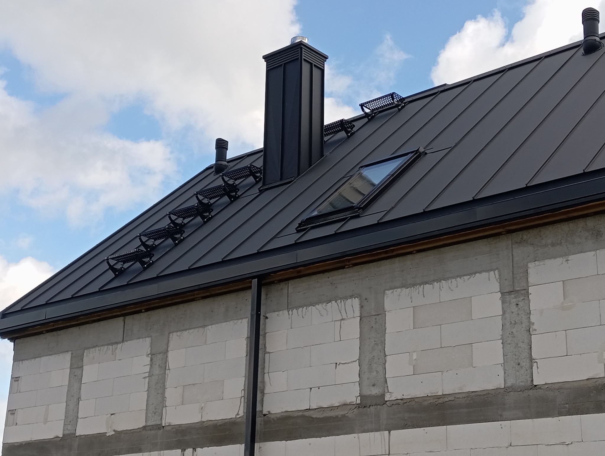 Czarny dach z blachy na domu w trakcie budowy, widoczne stopnie dachowe, komin i okno dachowe. W tle błękitne niebo z chmurami.