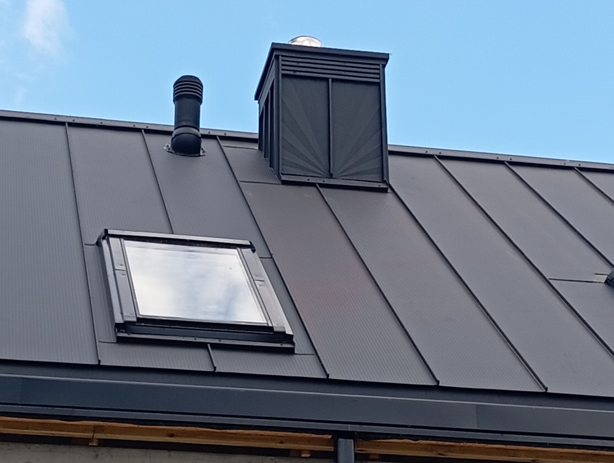 Szary dach z blachy na rąbek stojący z oknem dachowym, kominem wentylacyjnym i ozdobnym kominem. Widok na błękitne niebo.