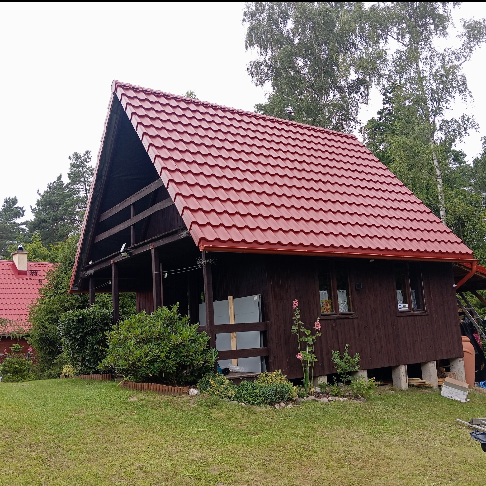 Ciemnobrązowy domek z czerwonym dachem z blachodachówki, otoczony zielenią. Widoczny fragment drugiego budynku z podobnym dachem.