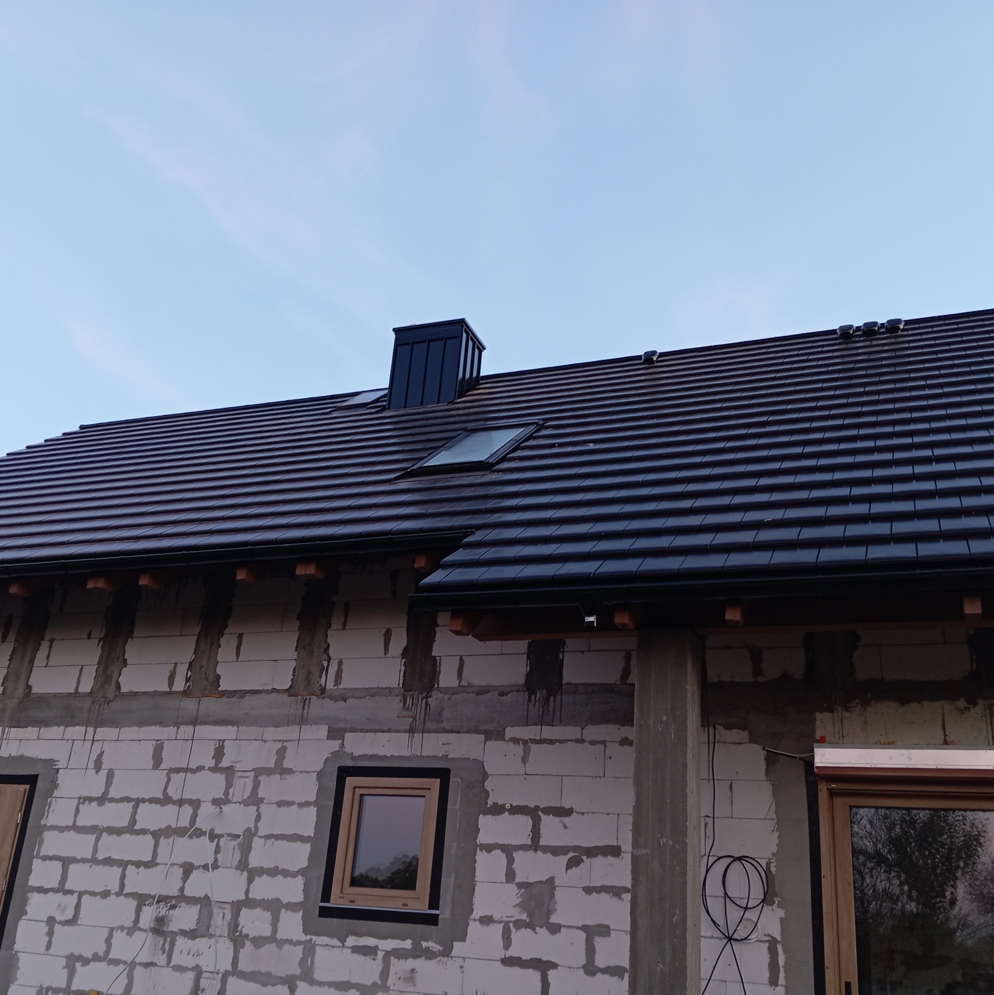 Nowy, ciemny dach z dachówką ceramiczną i oknem dachowym na budynku w trakcie budowy z elementami obróbek blacharskich i widocznym kominem.