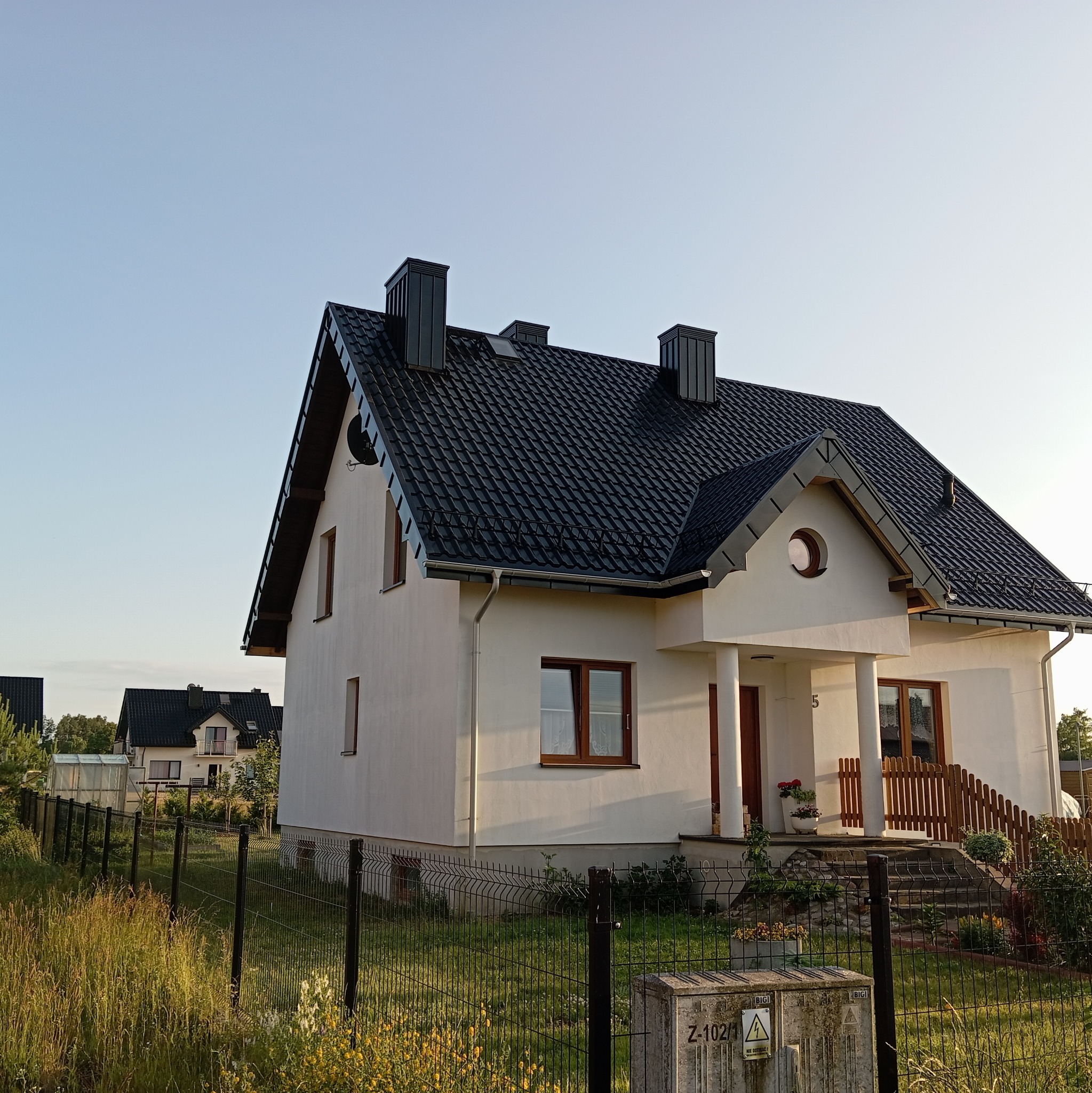 Biały dom jednorodzinny z czarnym, dwuspadowym dachem z blachodachówki, widoczny zza ogrodzenia. Na dachu trzy kominy. W tle drugi dom.