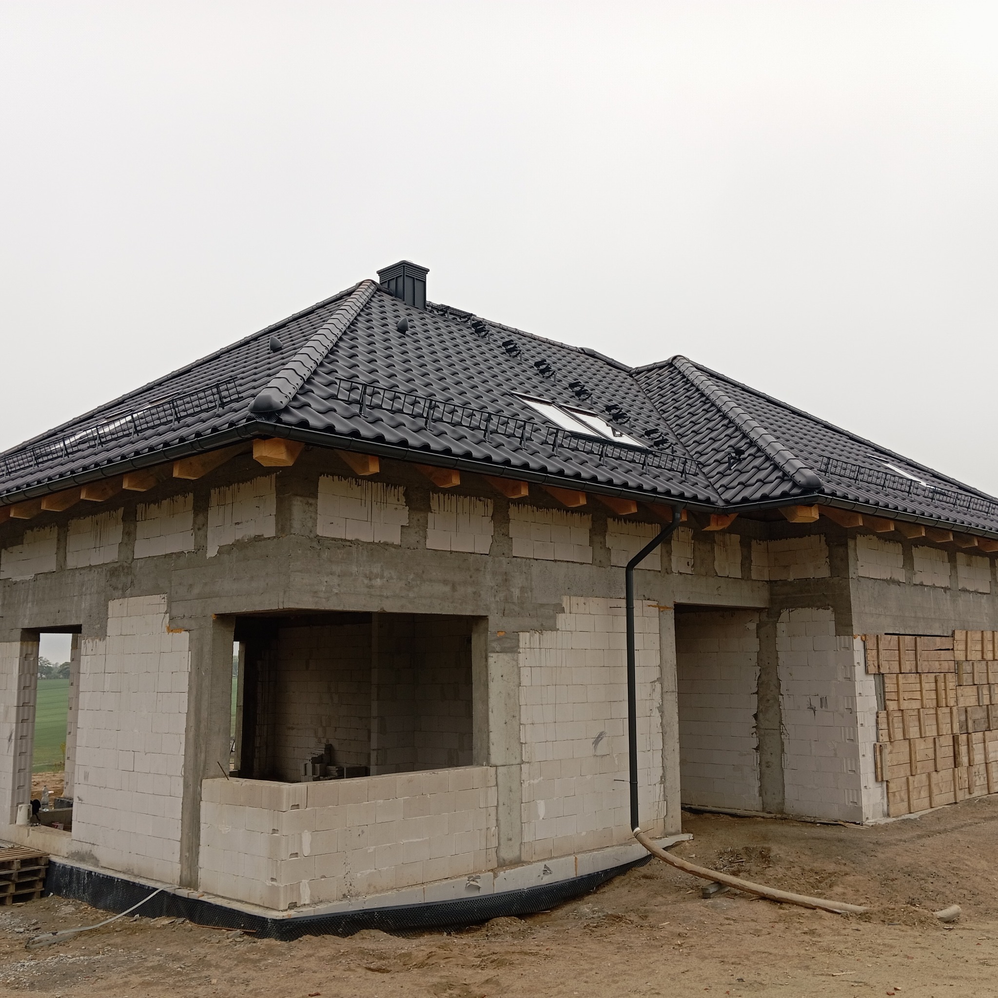 Surowy stan domu jednorodzinnego z czarnym dachem z dachówki ceramicznej, widoczne okna dachowe i rynny. Dom w trakcie budowy, ściany z białego bloczka.