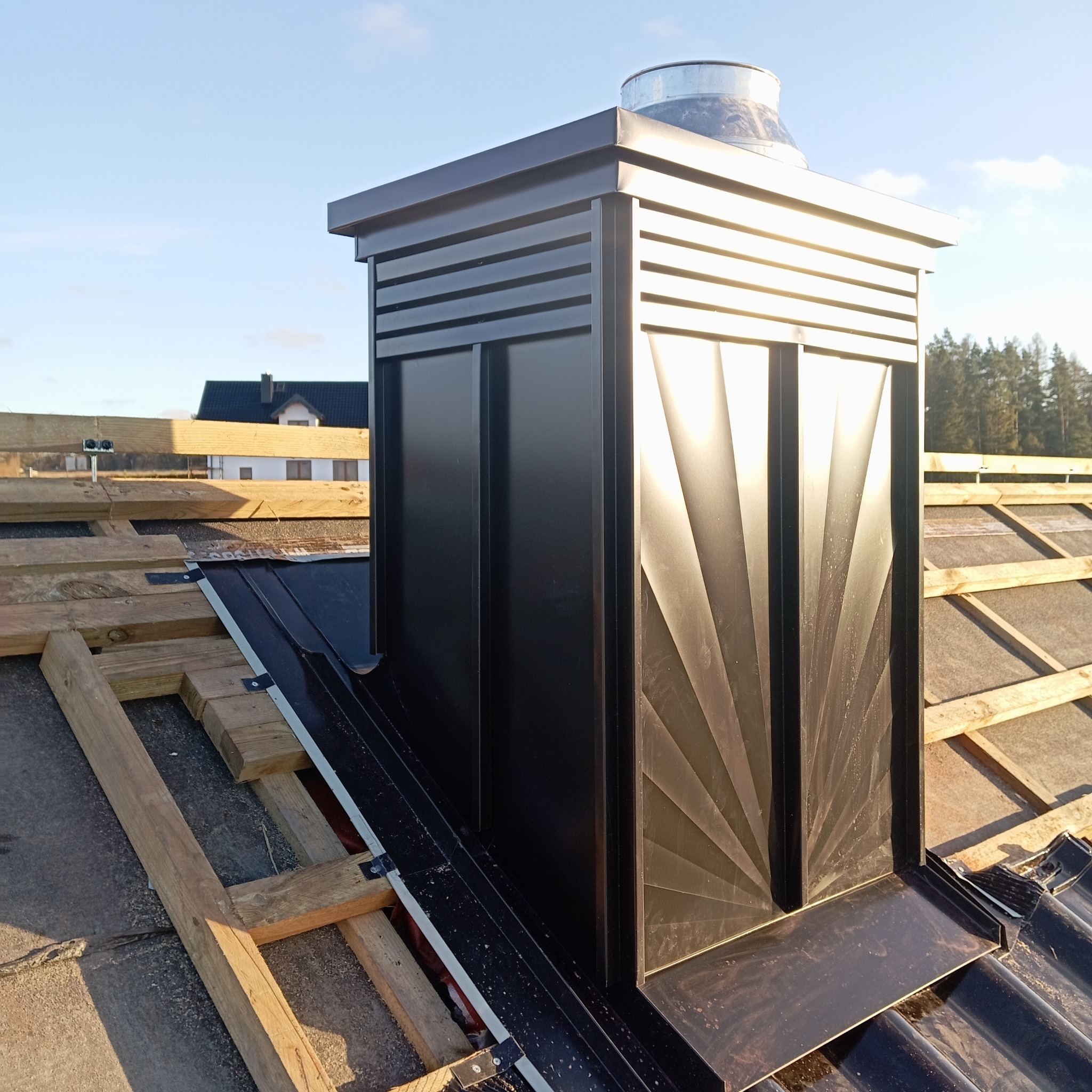Czarny komin wentylacyjny na dachu z drewnianą konstrukcją w tle, widok z góry, słoneczny dzień. Element dachu z blachy z charakterystycznym wzorem.
