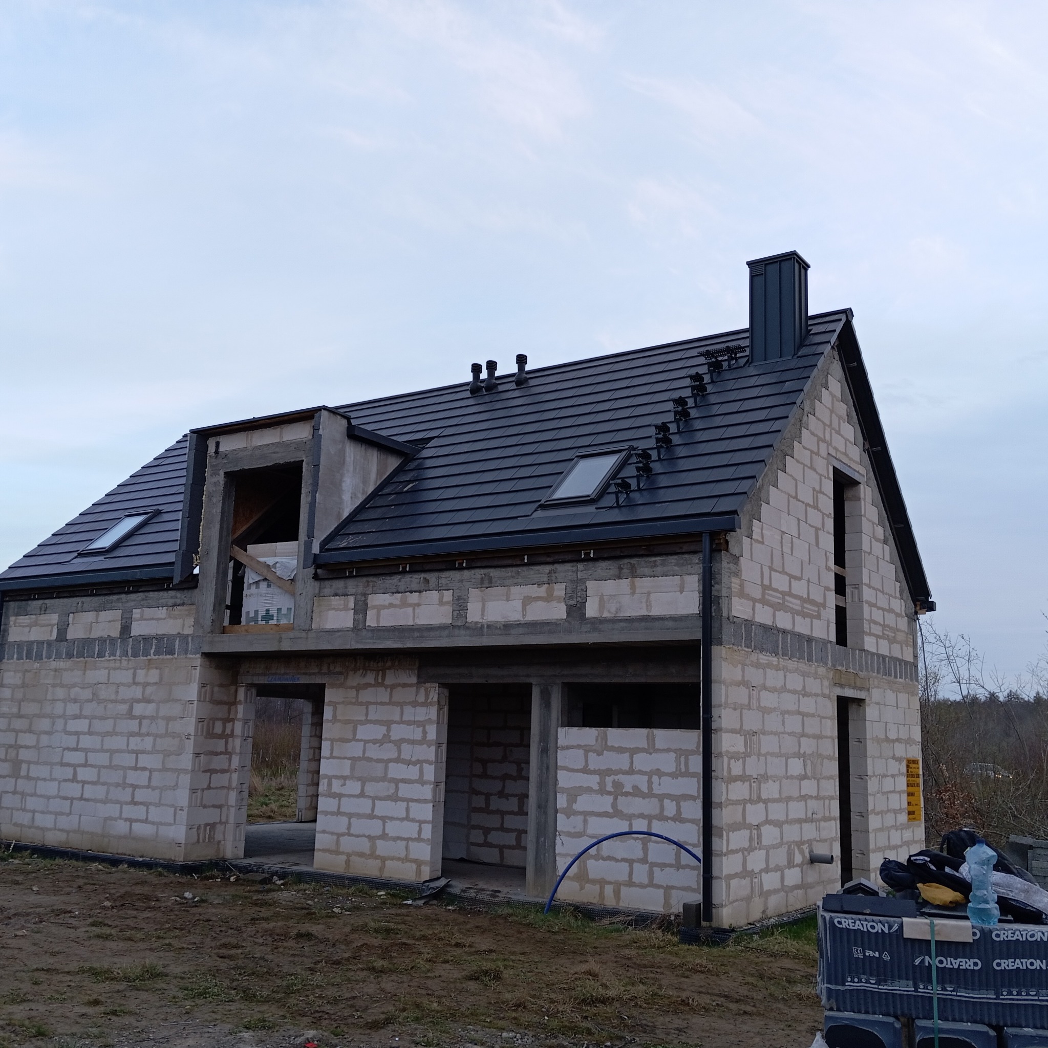Dom w trakcie budowy z czarnym dachem z dachówki ceramicznej Creaton, widoczne okna dachowe i komin. Ściany z białych bloczków, stan surowy otwarty.