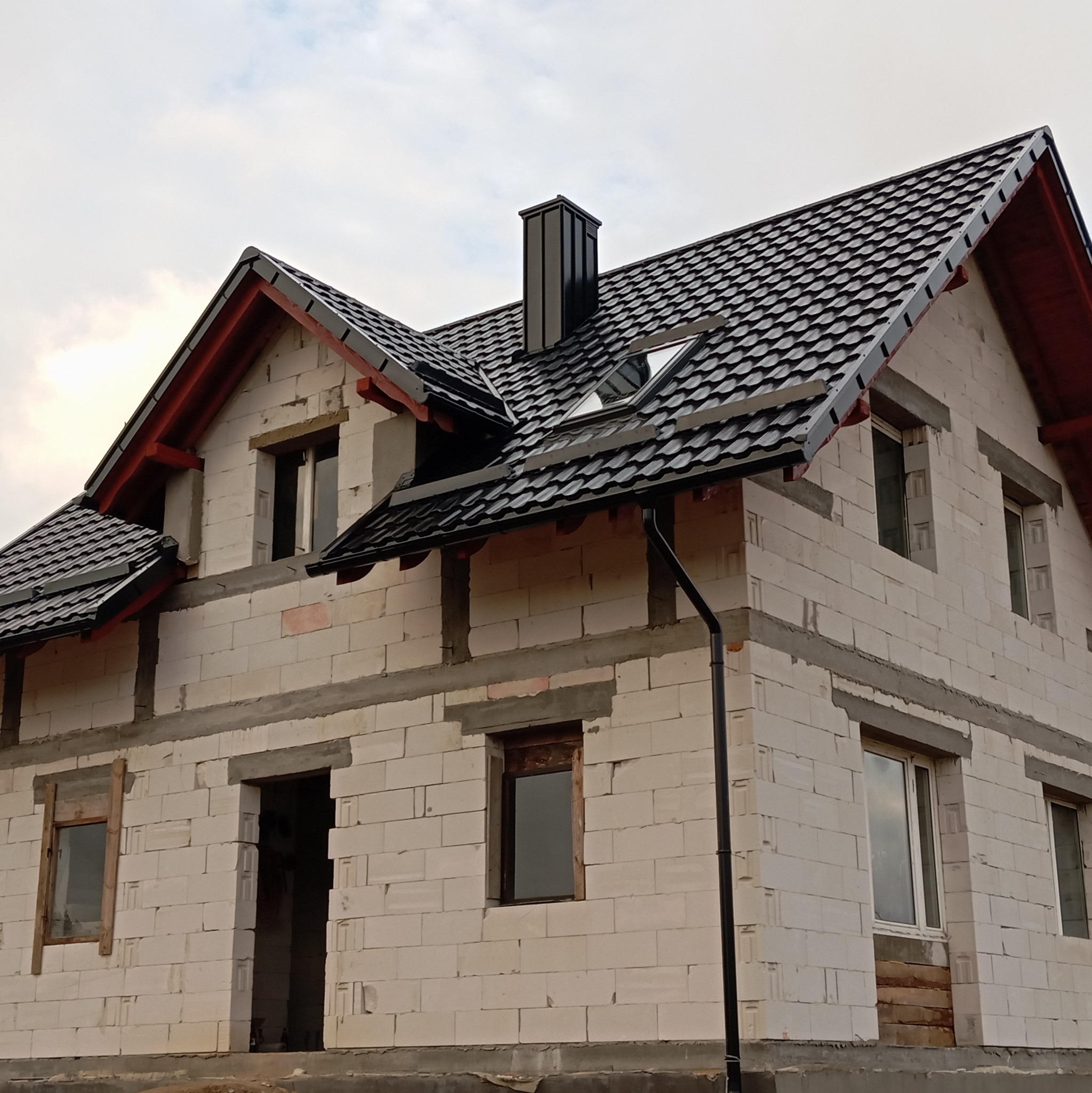 Dom w trakcie budowy z czarnym dachem z dachówki, widoczne rynny i komin. Okna częściowo zamontowane, ściany z białych bloczków.