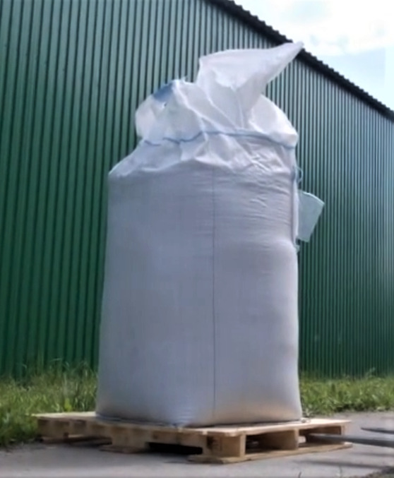 Wypełniony biały BIGBAG na drewnianej palecie, na tle zielonej, blaszanej ściany magazynu.