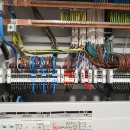 Szczegółowe ujęcie wnętrza szafy elektrycznej z licznymi przewodami w różnych kolorach, podłączonymi do listew zaciskowych i szyn zbiorczych, z etykietami na przewodach i modułami zabezpieczającymi...